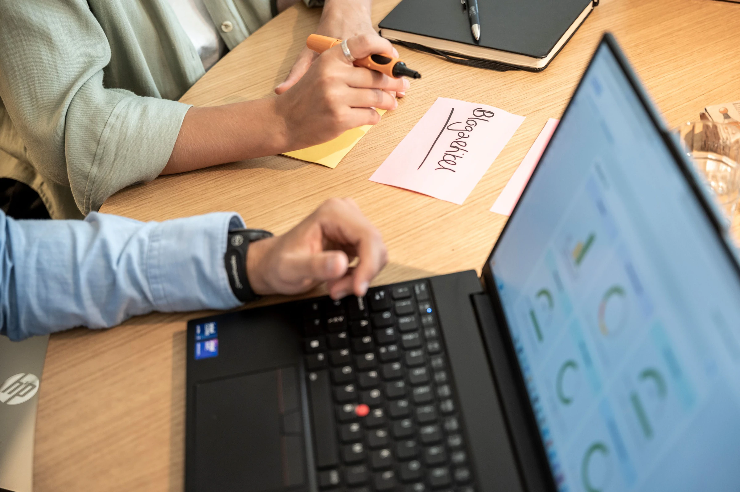 Zwei Personen am Tisch: Eine schreibt Karten für einen Workshop, die andere arbeitet am Laptop an einer Auswertung