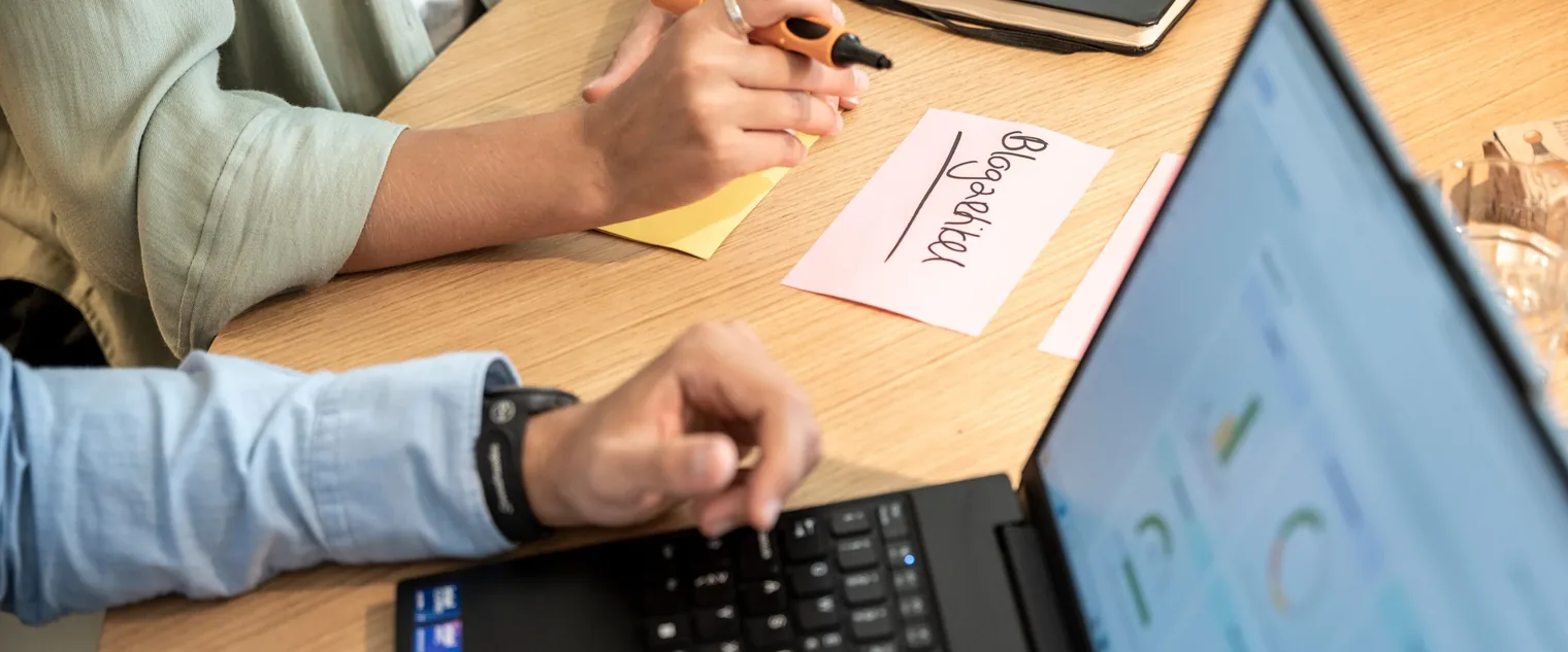 Zwei Personen arbeiten an einem Holztisch mit Laptop, Notizzettel und Marker